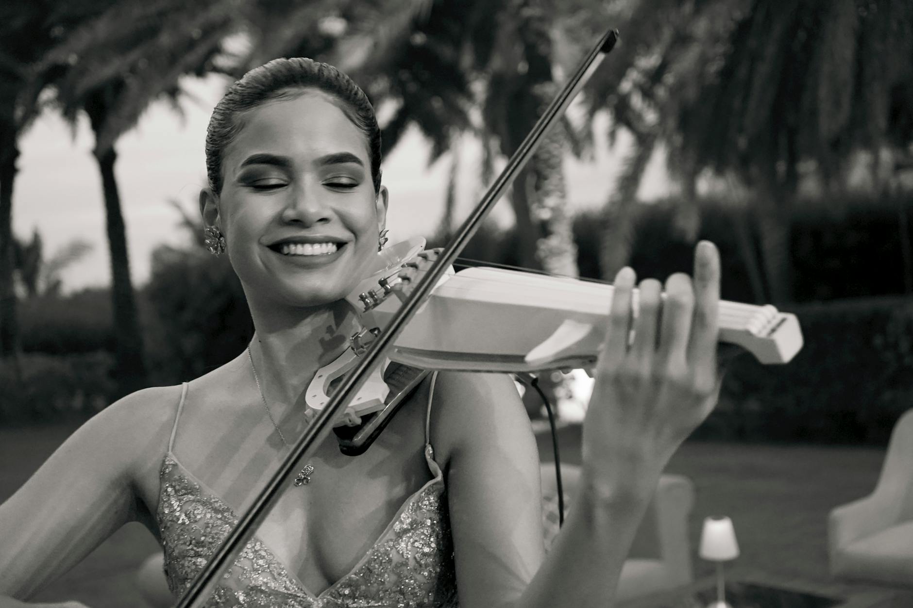 Smiling woman playing violin outdoors. Black and white image captures musical passion and elegance.